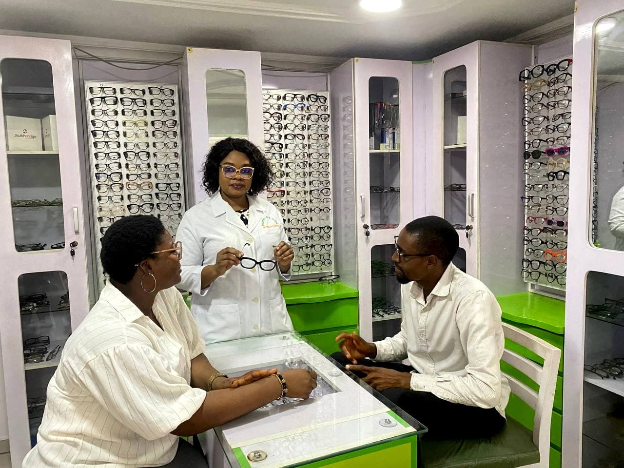 An optometrist holding up a pair of spectacles for a seated patient at the dispensing counter, with walls of frame cabinets on both sides.