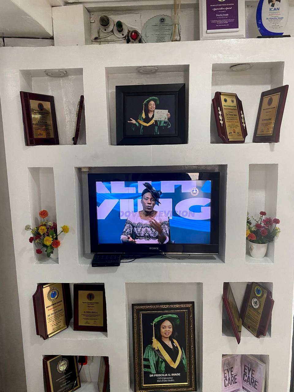 The clinic's wall of framed certificates, awards and photographs, arranged in white alcoves around a wall-mounted screen.