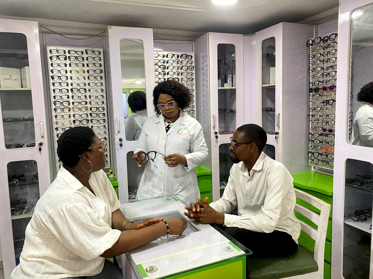 Two members of staff helping a patient choose spectacles at the dispensing counter, surrounded by lit frame displays.