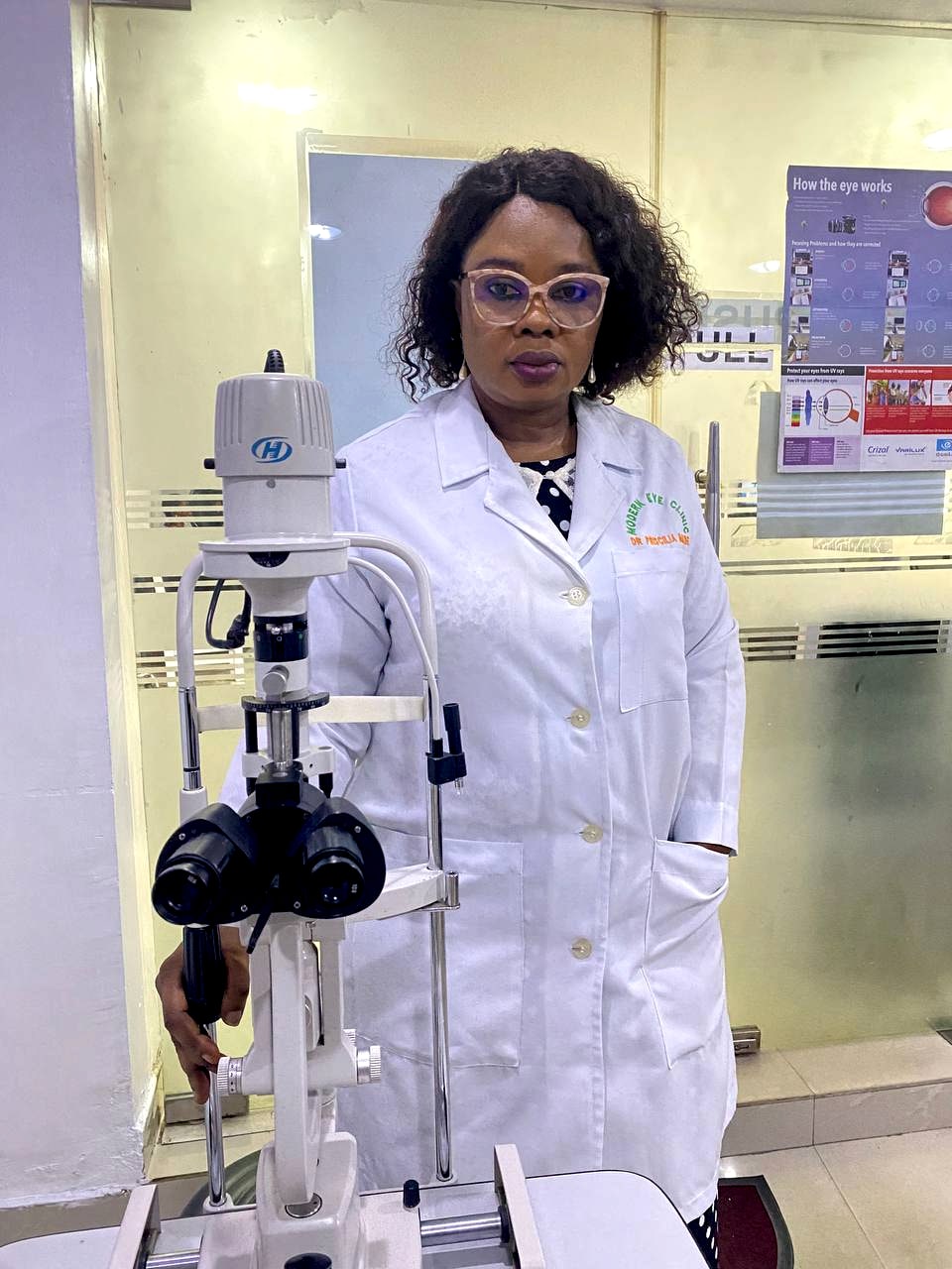 Dr. Priscilia Imade in a white coat, standing beside the slit lamp in the examination room. A diagram headed How the eye works is on the wall behind her.