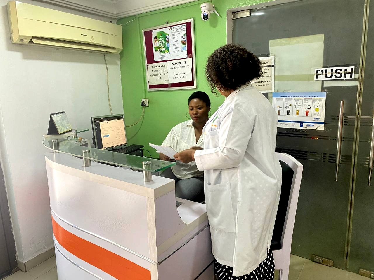 Two members of staff at the clinic's reception counter, one in a white coat, against the green wall of the front room.