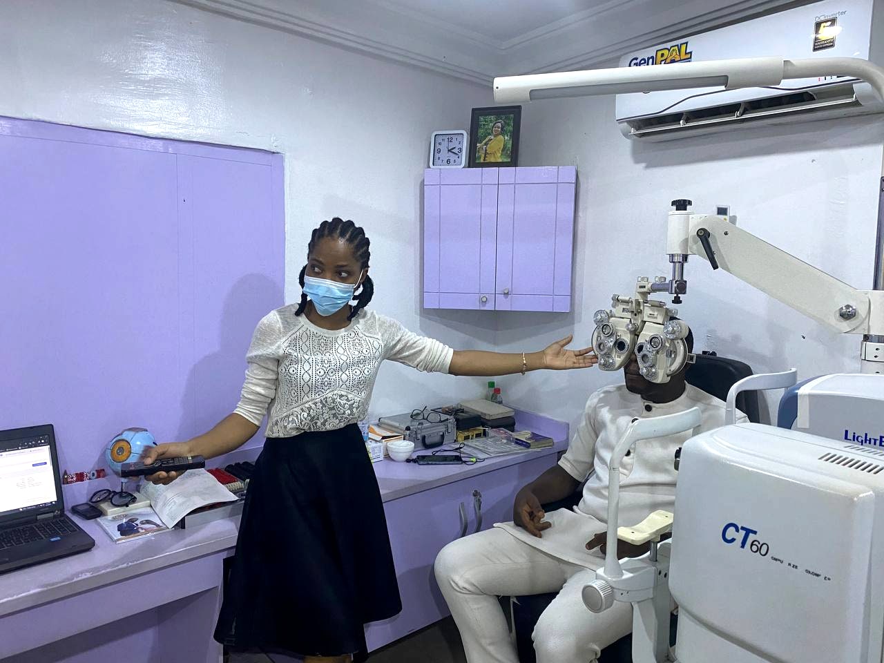 A clinician reaching for the phoropter arm beside a seated patient, with a laptop and tonometer on the bench behind her.