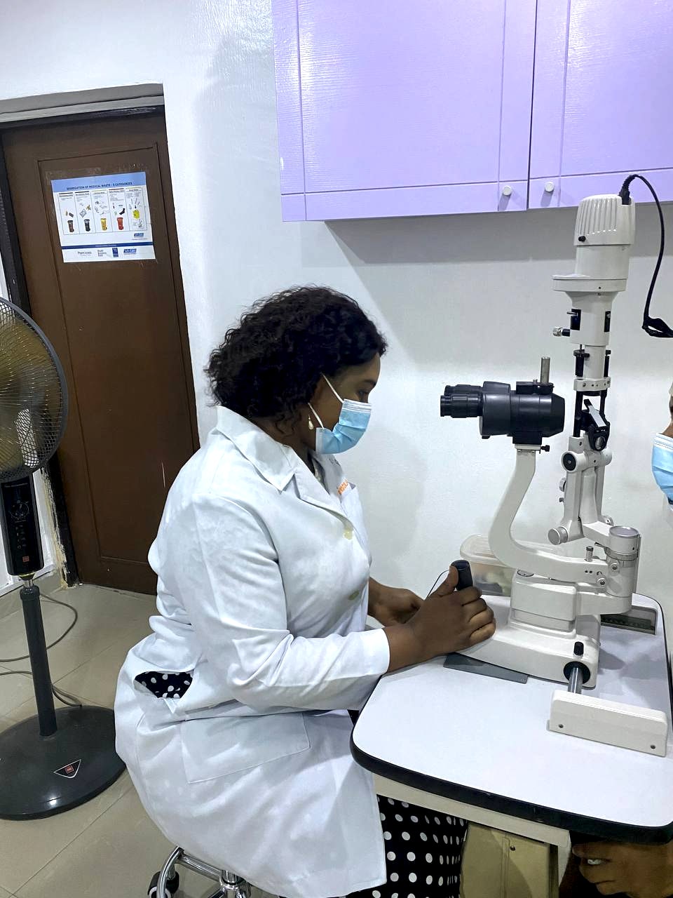 An optometrist seated at a slit lamp biomicroscope, hands on its controls, examining the front of a patient's eye.