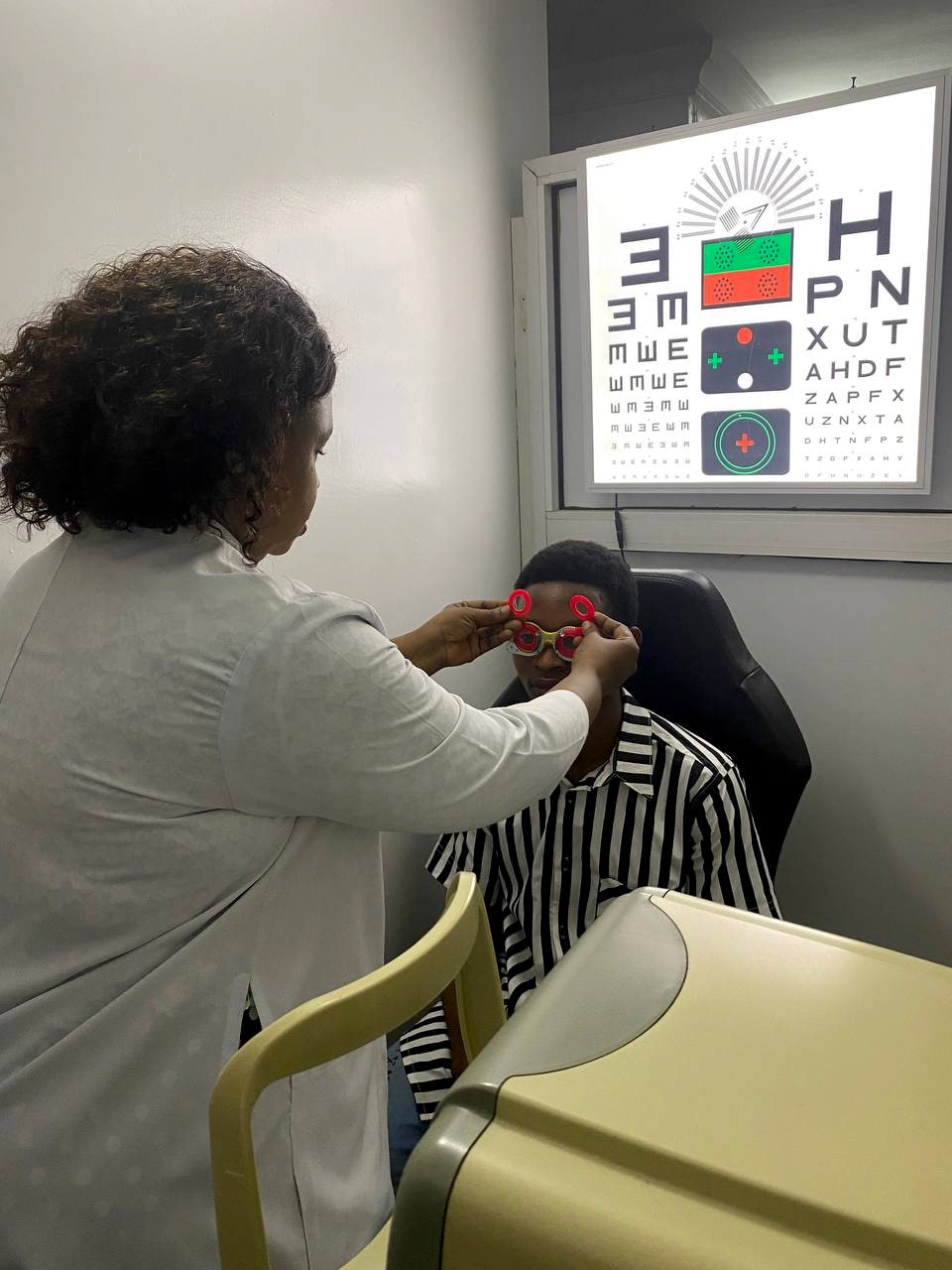 An optometrist standing behind a seated patient, fitting a red trial frame to their face. An illuminated letter chart is on the wall in front of them.